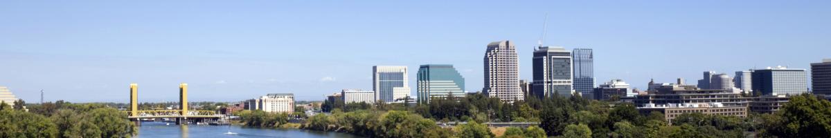 green trees line both banks of the sacramento river. the yellow tower bridge crosses the blue water leading to the tall buildings of the sacrament skyline. the scene is set against a clear blue sky.