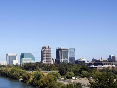 green trees line both banks of the sacramento river. the yellow tower bridge crosses the blue water leading to the tall buildings of the sacrament skyline. the scene is set against a clear blue sky.